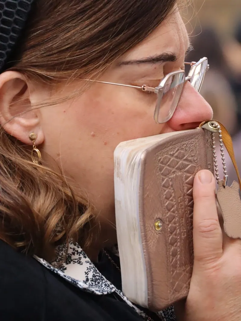 אישה במשקפיים מצמידה לשפתיה ספר תפילה קטן בכריכה מעוטרת, בעיניים עצומות כמעט.