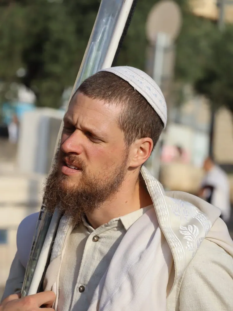 גבר מזוקן בכיפה סרוגה לבנה ובטלית, נושא על כתפו לולב עטוף בנרתיק פלסטיק.