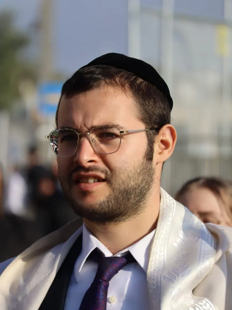 צעיר בכיפה שחורה, משקפיים ועניבה, עטוף בטלית לבנה, ברחוב בשעת אור רכה.