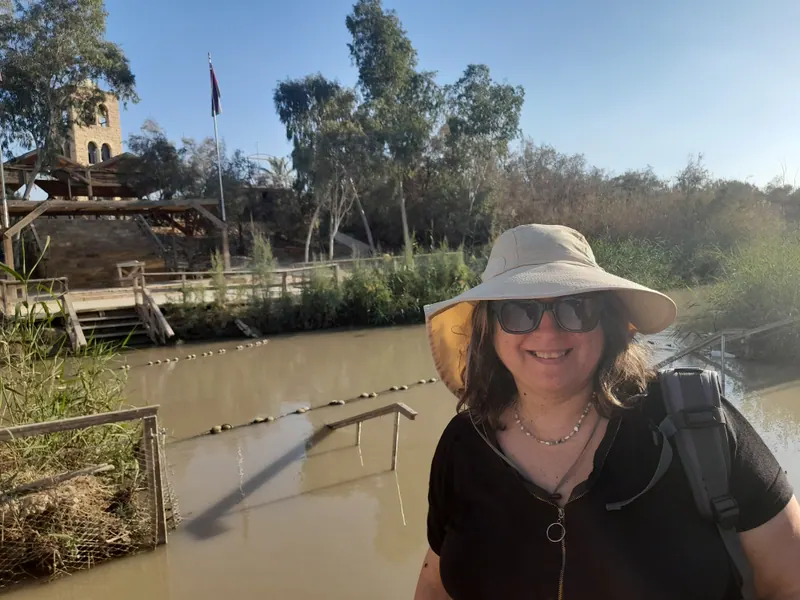 רחל אפרסמון מחייכת, בכובע רחב שוליים, משקפי שמש ותרמיל, על גדת נהר הירדן; מאחוריה חבל חוצה את המים, רציף עץ, קנים ועצים.