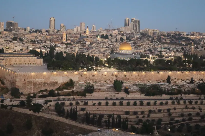העיר העתיקה בשעת בין ערביים במבט מהר הזיתים: החומה המזרחית מוארת, כיפת הסלע במרכז ומדרונות מדורגים מתחתיה.