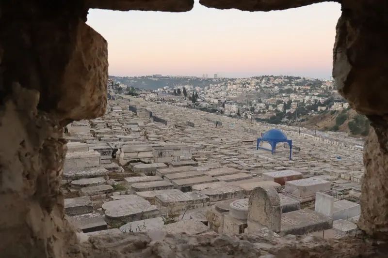 מבט דרך פתח באבן אל בית הקברות היהודי בהר הזיתים, עם מבנה קבר בעל כיפה כחולה, ומעבר לו שכונות על הגבעות.