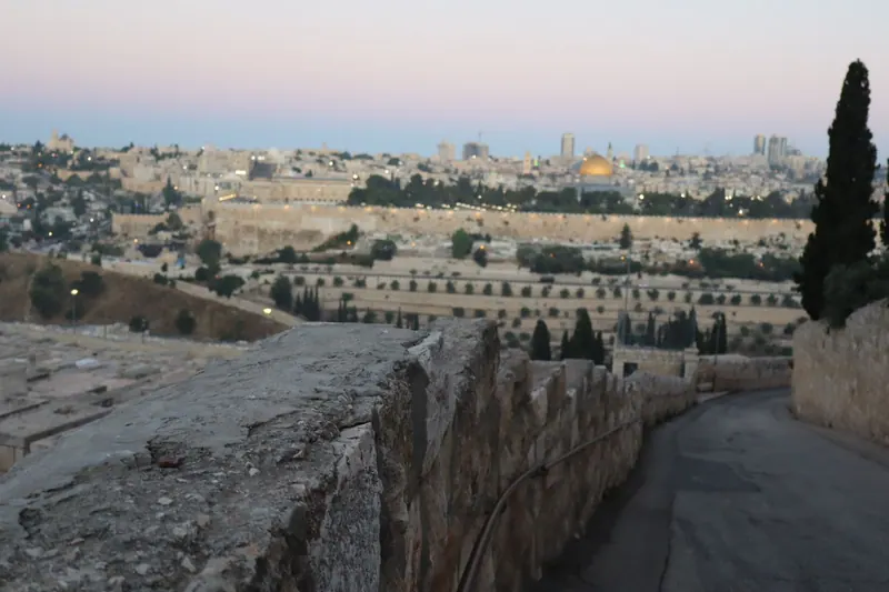 דרך יורדת לאורך חומת אבן בהר הזיתים בדמדומים, ומולה העיר העתיקה עם כיפת הסלע המוזהבת.