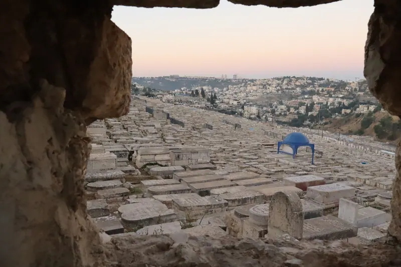 מצבות אבן צפופות על מדרון, במבט מבעד לפתח בסלע, תחת שמי שקיעה ורודים.