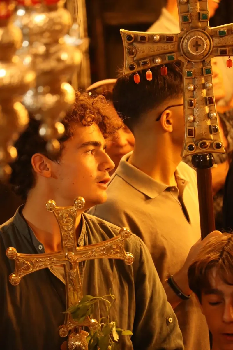 צעירים נושאים צלבי תהלוכה מוזהבים משובצי אבנים בטקס כנסייה, תחת מנורות כסף תלויות.