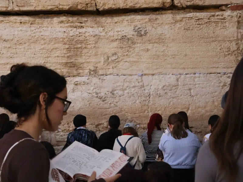 עזרת הנשים בכותל המערבי: נשים מתפללות מול אבני הכותל, ובקדמת התמונה אישה צעירה קוראת בסידור.