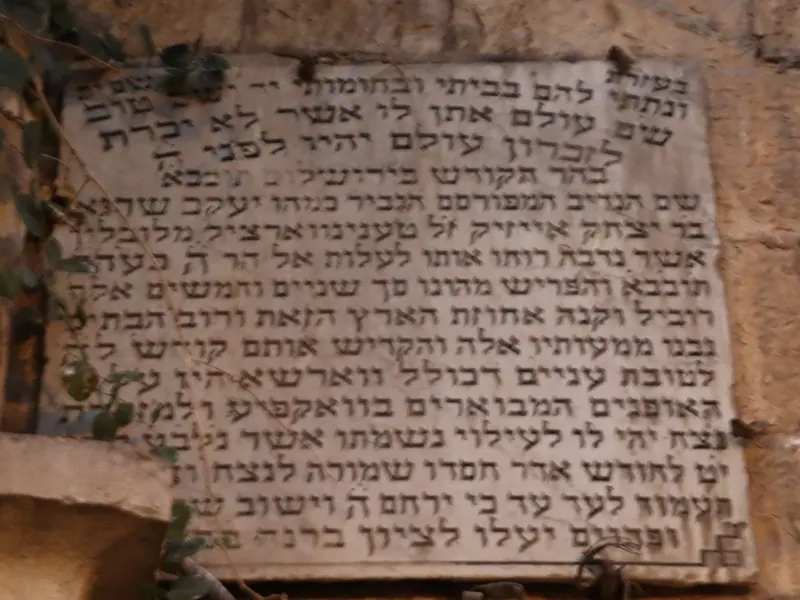 לוח אבן ישן חקוק בכתב עברי צפוף, קבוע בקיר ומוקף צמחייה.