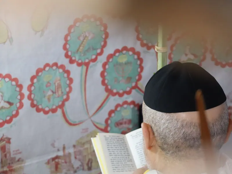 גבר בכיפה שחורה קורא בספר תפילה מול בד סוכה מעוטר, מצולם מאחור.