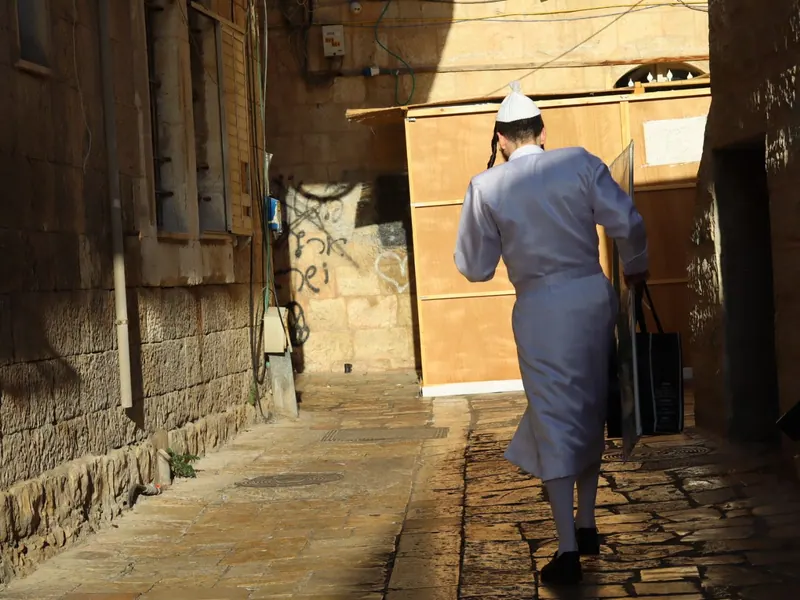 גבר בחלוק בהיר ובכיפה לבנה הולך בסמטה מרוצפת אבן אל עבר סוכת עץ מוארת, מצולם מאחור.