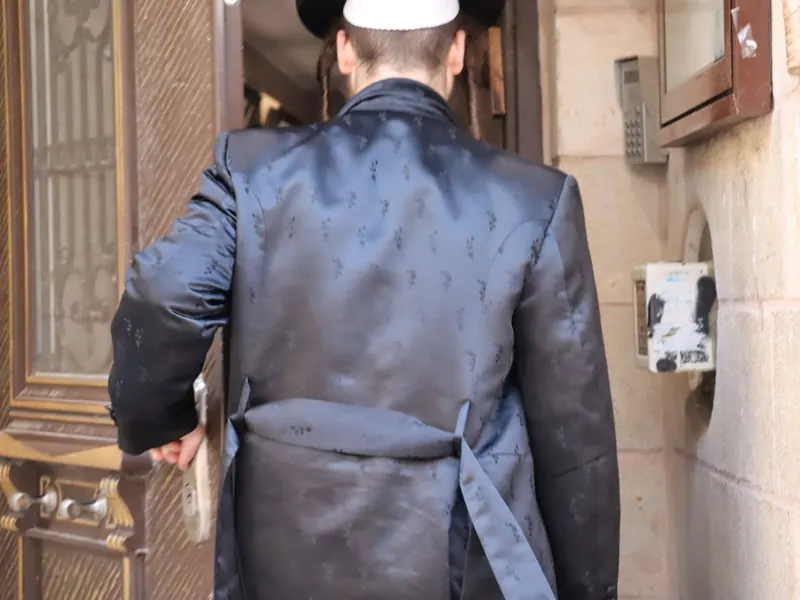 גבו של חסיד בקפוטה משי כהה רקומה וחגורת גארטל, נכנס בפתח עץ בבניין אבן.