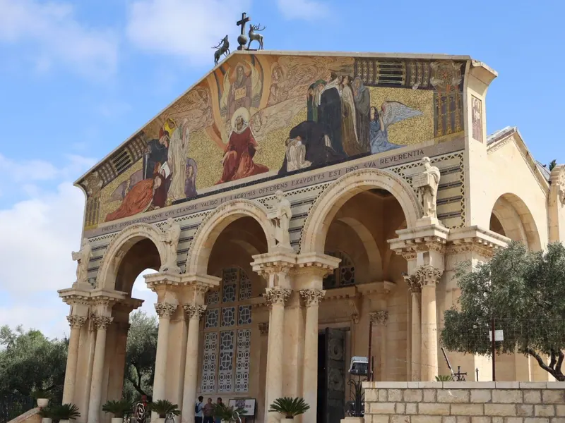 חזית כנסיית כל העמים בגת שמנים: פסיפס מוזהב מעל שורת קשתות ועמודים, ועצי זית לצדה.