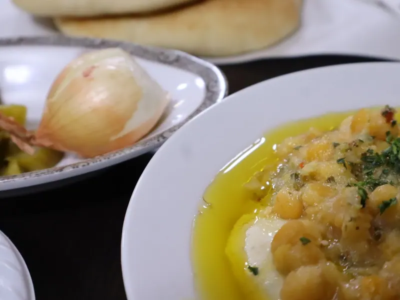 צלחת חומוס עם גרגרים שלמים ושמן זית, ולידה בצל חצוי, חמוצים ופיתות.