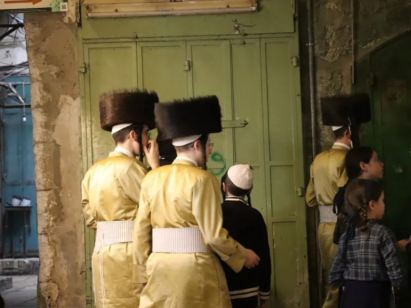חסידים בגלימות מפוספסות זהובות ובשטריימלים עומדים בסמטה בעיר העתיקה מול דלתות ירוקות, לצד ילדים.