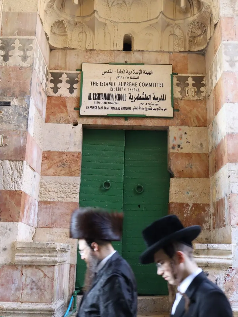 פתח המדרסה הטשתמריה בעיר העתיקה: דלת ירוקה, אבן אדומה ולבנה ושלט מעליה, ושני גברים חרדים חולפים לפניה.