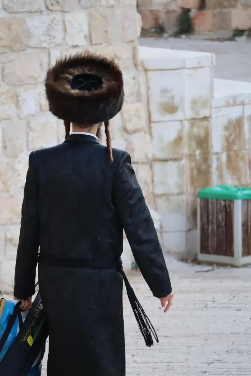 נער חסידי בשטריימל וקפוטה שחורה, פאות ארוכות וחגורת גארטל, הולך בגבו למצלמה לאורך קיר אבן.