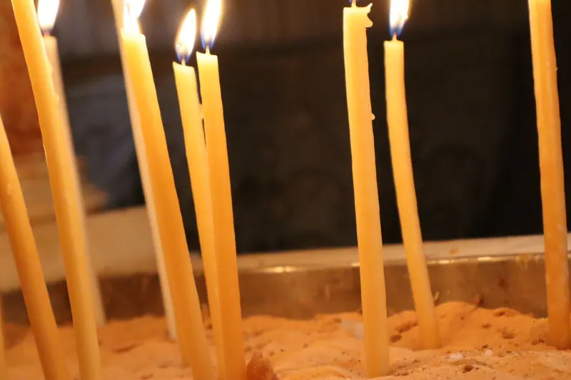נרות שעווה דקים וארוכים בוערים, נעוצים בחול בתוך מגש מתכת באפלולית.