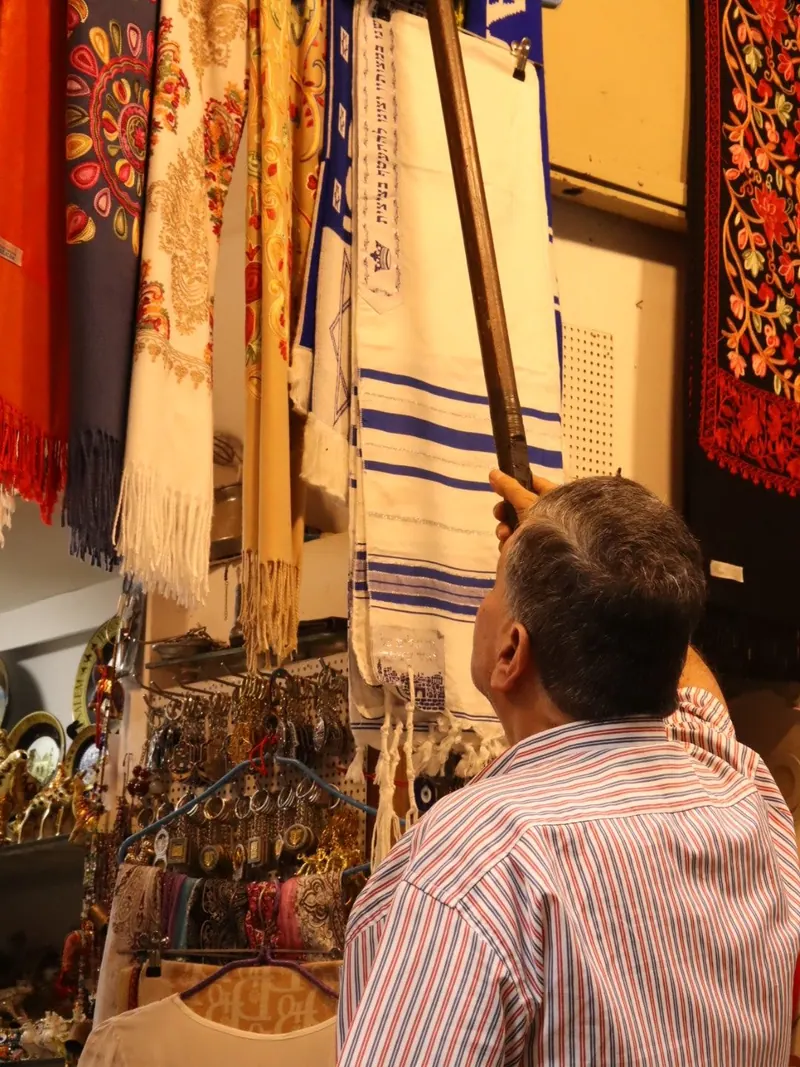 מוכר בחנות מוריד במוט טלית לבנה עם פסים כחולים, בין צעיפים רקומים ומחזיקי מפתחות.