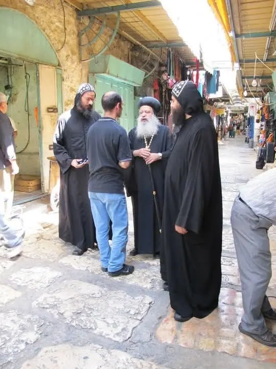 אנשי דת קופטים בגלימות שחורות ובכיסויי ראש רקומים משוחחים עם אדם בסמטת שוק מקורה בעיר העתיקה.