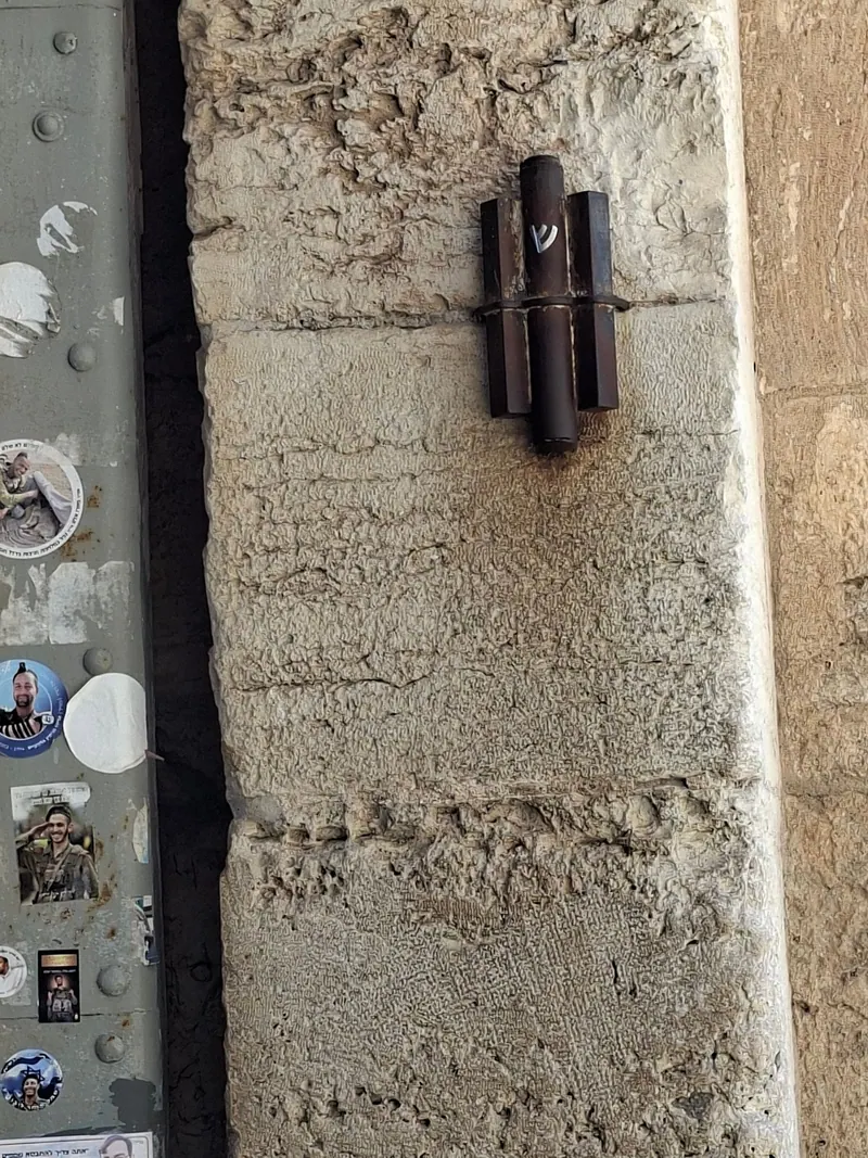מזוזה בבית מתכת כהה על עמוד אבן גדול של שער, ולצדה דלת מתכת מכוסה מדבקות.