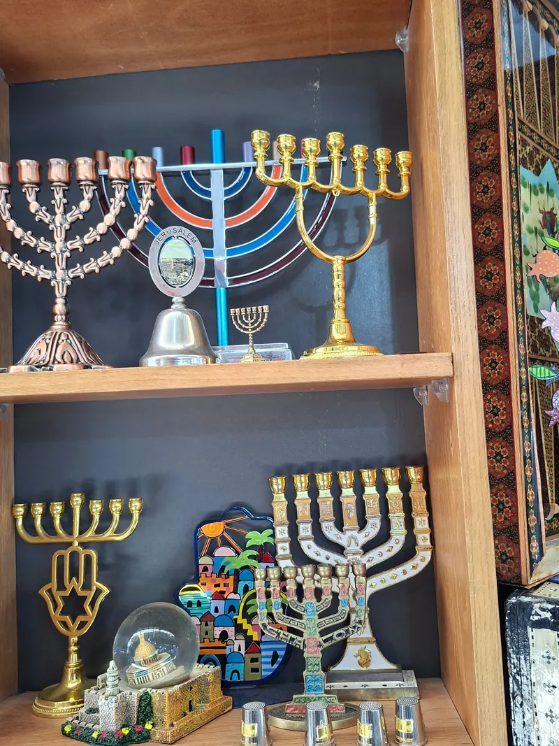 מדפים של מנורות שבעה קנים בנחושת, בפליז ובצבעים, לצד כדור שלג עם כיפת הסלע ומזכרות מירושלים.