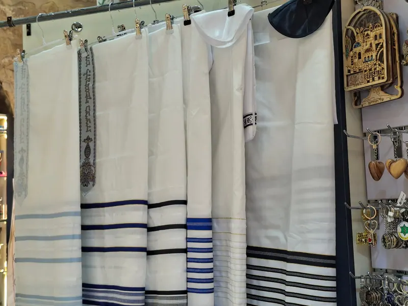 טליתות לבנות עם פסים כחולים ושחורים תלויות על קולבים בחנות, לצד מחזיקי מפתחות ומגנטים מעץ זית.
