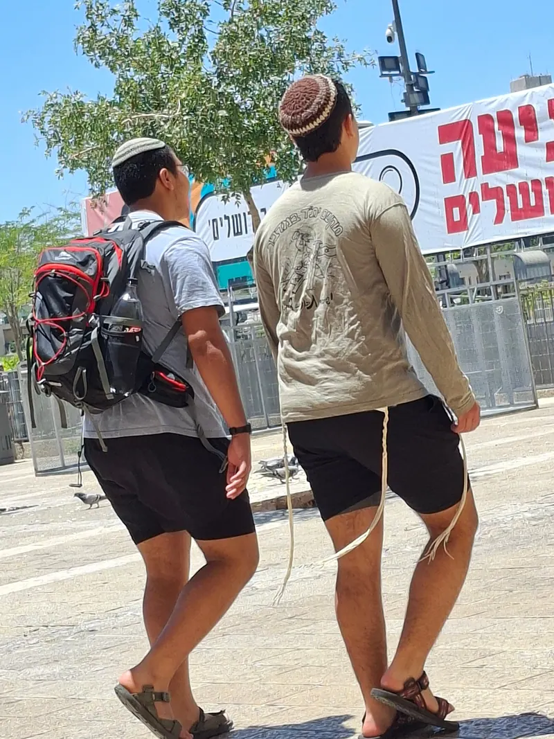 שני צעירים חובשי כיפות סרוגות וציציות משתלשלות הולכים ברחבה, מול שלט גדול בעברית.