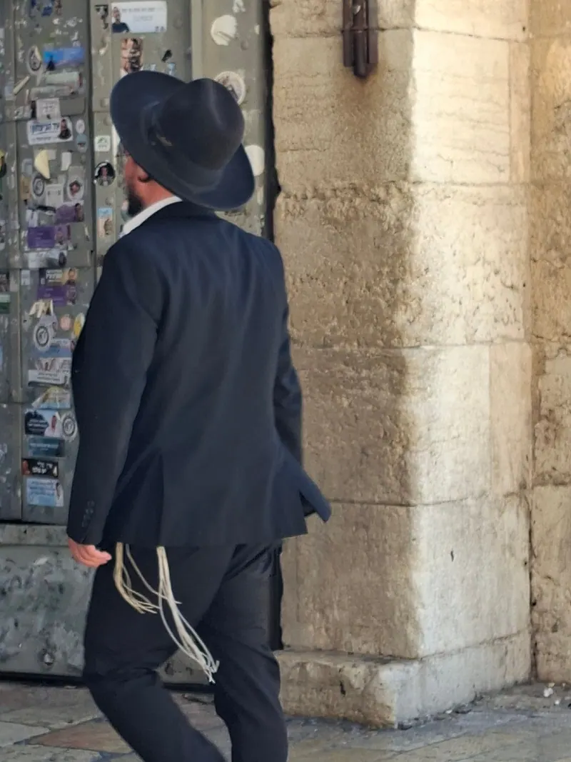 גבר חרדי בכובע שחור ובחליפה, ציציותיו משתלשלות, עובר ליד שער אבן שעליו מזוזה.
