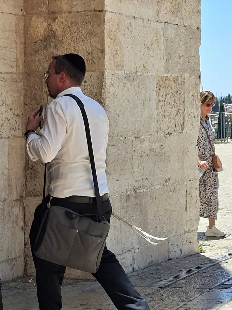 גבר בכיפה שחורה ובציציות ניגש אל עמוד האבן של שער, ומאחור אישה עומדת באור השמש.