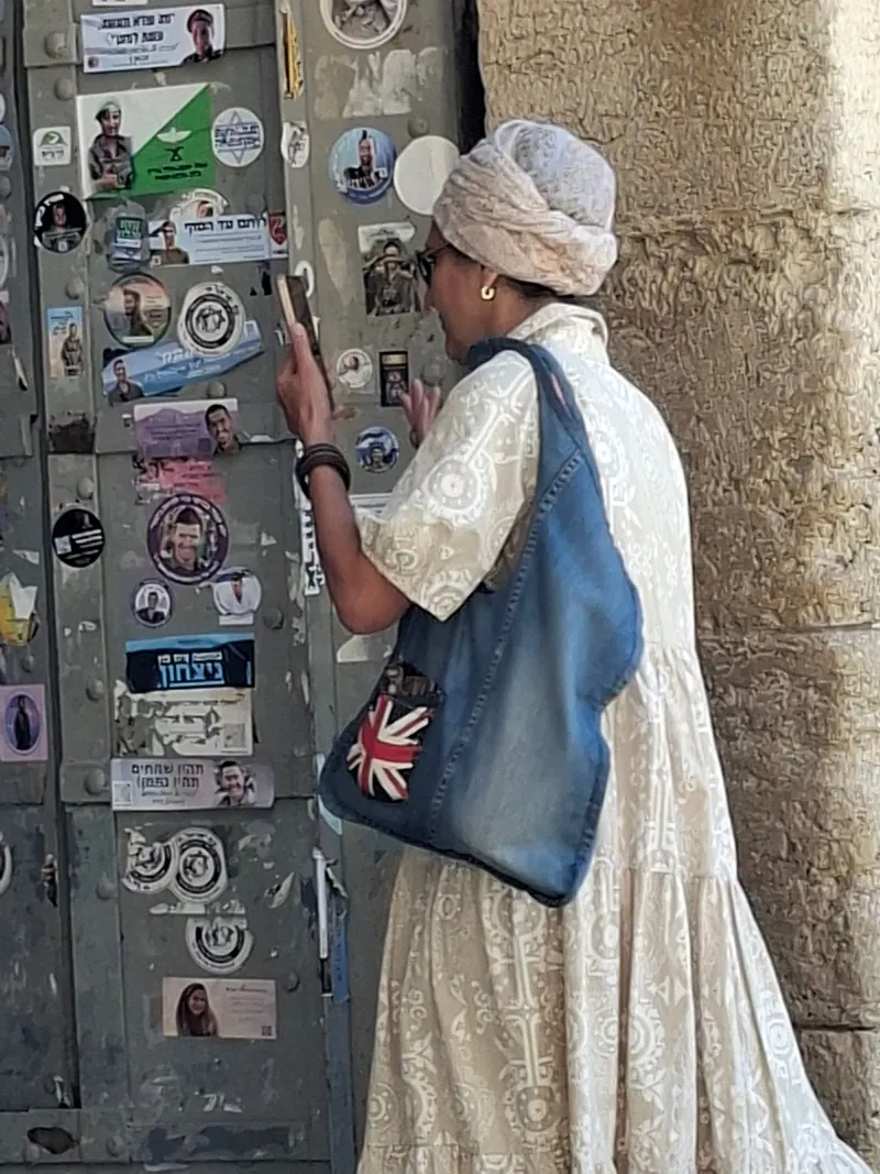 אישה בטורבן ובשמלה לבנה מצלמת דלת ברזל המכוסה מדבקות עם פניהם של חיילים, ליד קיר אבן.