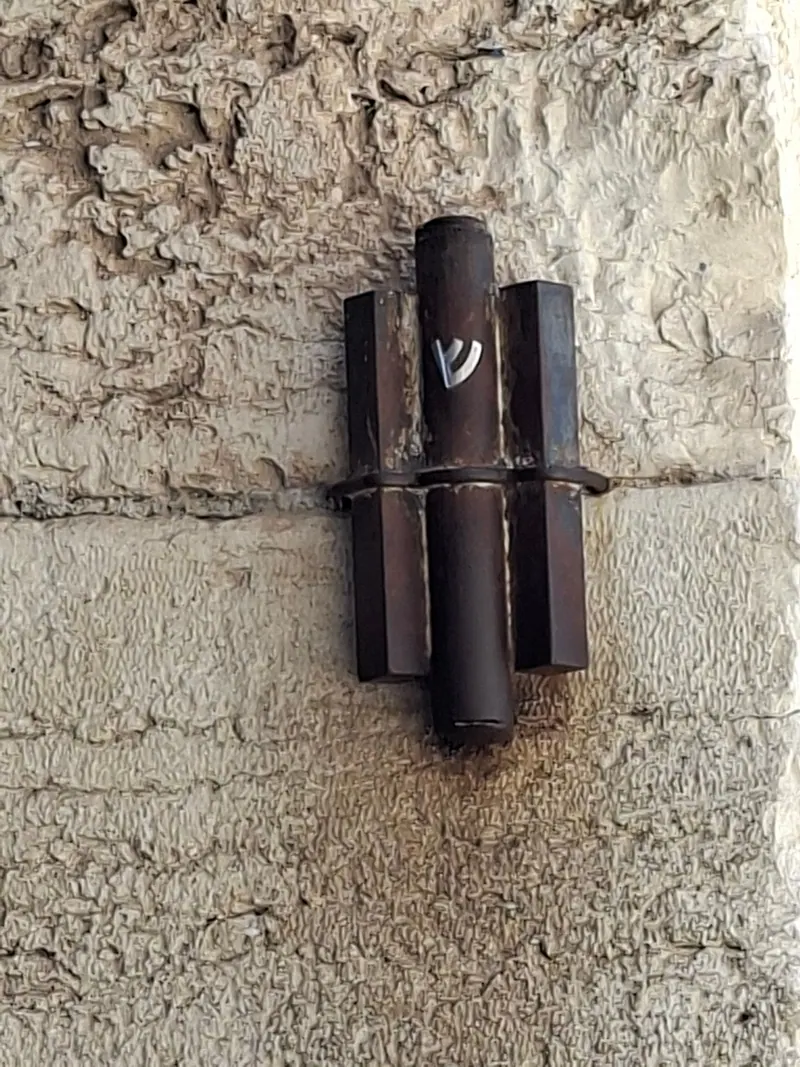 מזוזת מתכת כהה ועליה האות שי"ן, קבועה על קיר אבן מחוספס.