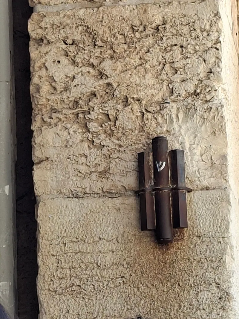 מזוזה בבית מתכת כהה, עליה האות ש, קבועה על משקוף אבן מסותתת.