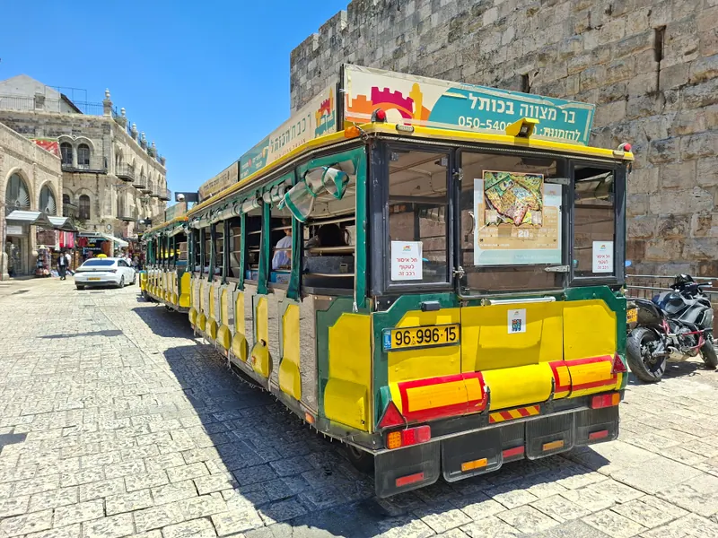 רכבת תיירים צהובה-ירוקה עם שלט "בר מצווה בכותל" חונה ברחוב מרוצף ליד חומת אבן גבוהה.