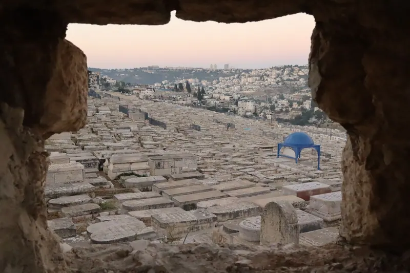 מבט מבעד לפתח באבן אל בית קברות יהודי גדול במדרון, עם כיפה כחולה על עמודים ושכונות ברקע, בשקיעה.