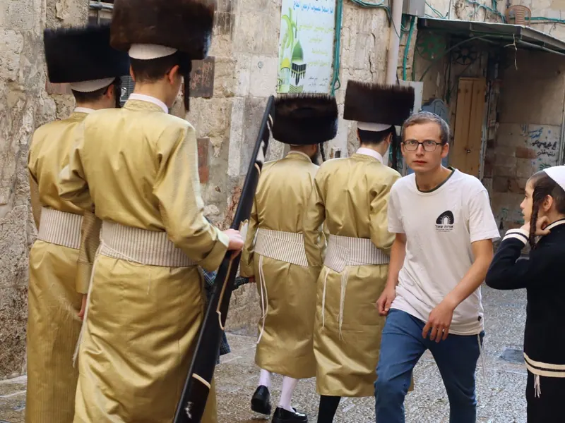 חסידים בגלימות מפוספסות זהובות ובשטריימלים צועדים בסמטה בעיר העתיקה, ולצדם צעיר בחולצה לבנה וילד.