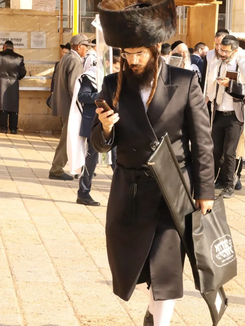 חסיד בשטריימל ובקפוטה שחורה, נושא תיק טלית ומביט בטלפון, ברחבת הכותל בין מתפללים נוספים.