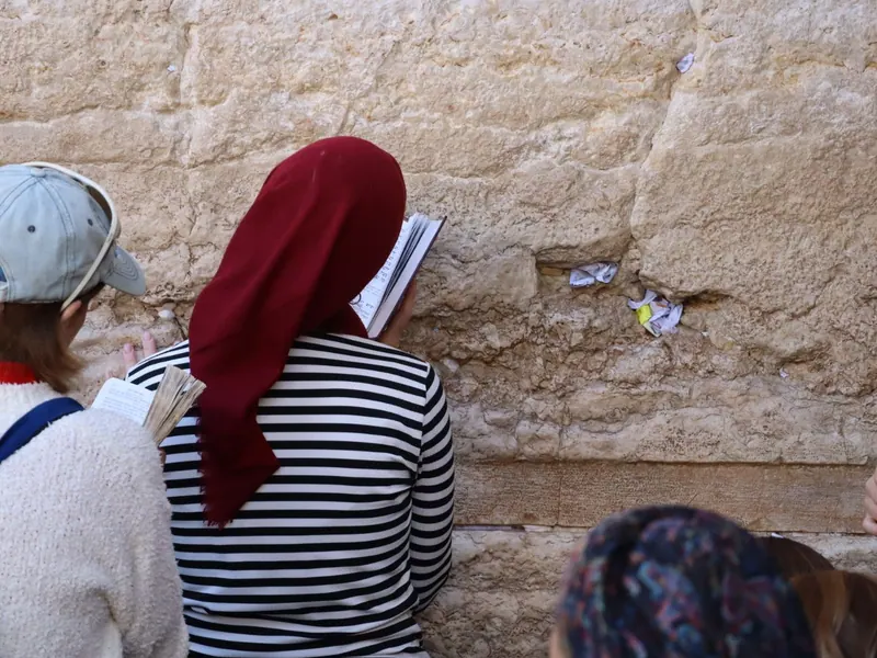 אישה במטפחת ראש אדומה וחולצת פסים קוראת בספר תפילה צמוד לאבני הכותל, מצולמת מאחור; פתקים תחובים בין האבנים.