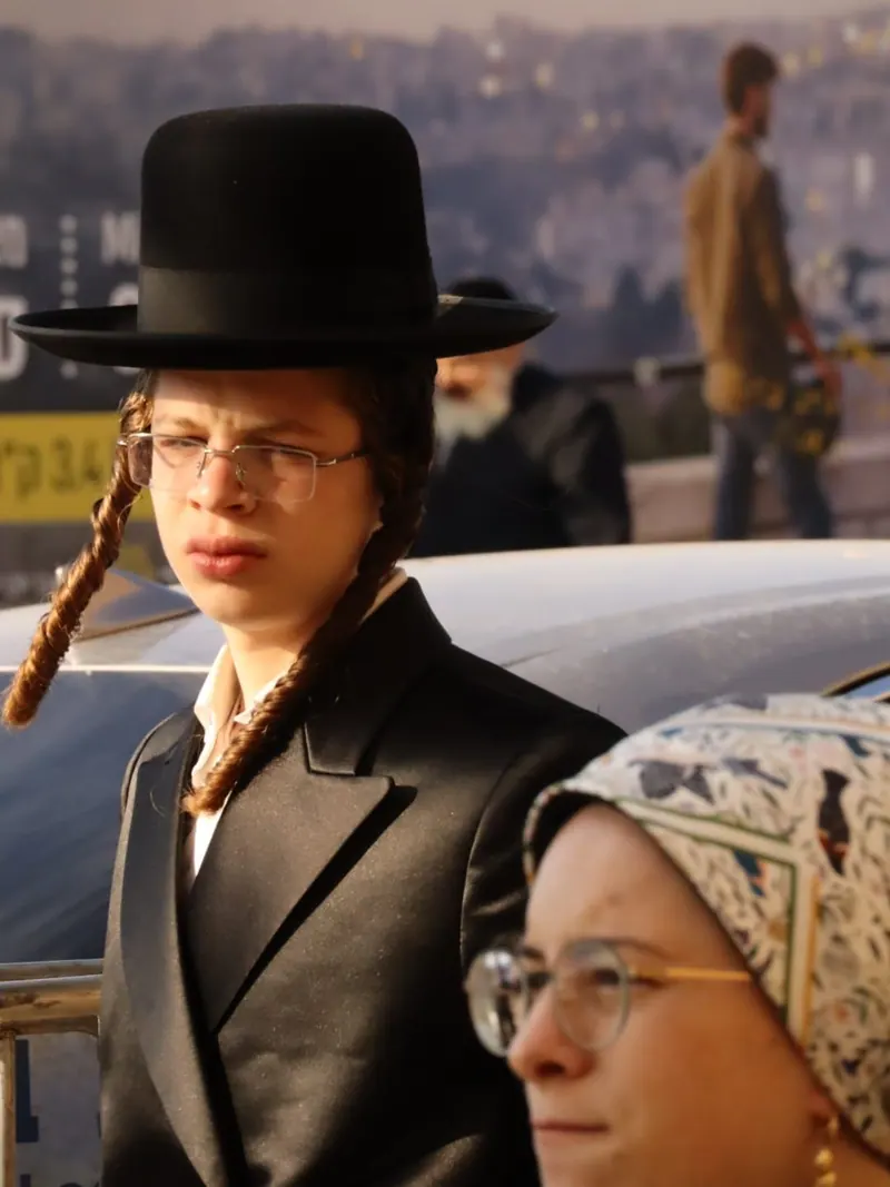 נער חסידי בכובע שחור רחב שוליים, משקפיים ופאות ארוכות מסולסלות, ולידו אישה במטפחת ראש מעוטרת.