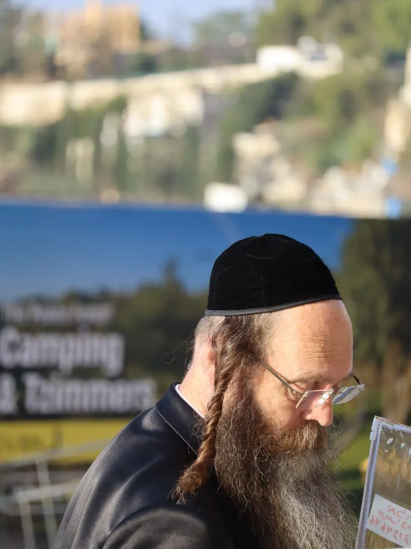 איש חרדי מזוקן בכיפת קטיפה שחורה ופאות ארוכות קלועות, מעיין בדף, ומאחוריו נוף מדרון ירוק ומים.