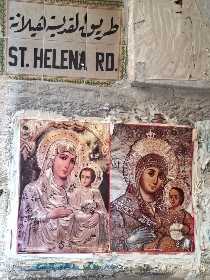 שלט רחוב בערבית ובאנגלית, ST. HELENA RD., ומתחתיו שתי כרזות של איקונות מרים והילד על קיר מסויד.
