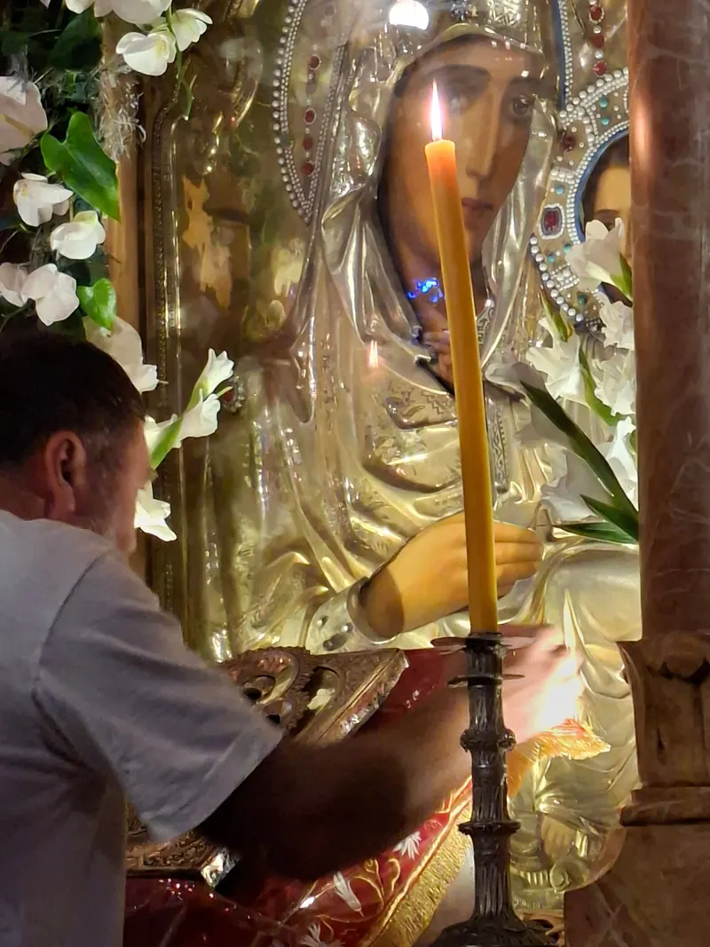 איש מושיט יד אל איקונין מכוסה כסף של מריה, מעוטר בפרחים לבנים, ולפניו נר שעווה דק דולק על פמוט.