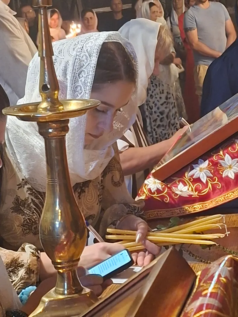 מתפללת בכיסוי ראש לבן מתחרה רוכנת אל איקונין ובידה נרות שעווה דקים, ומאחוריה קהל נשים בכיסויי ראש.
