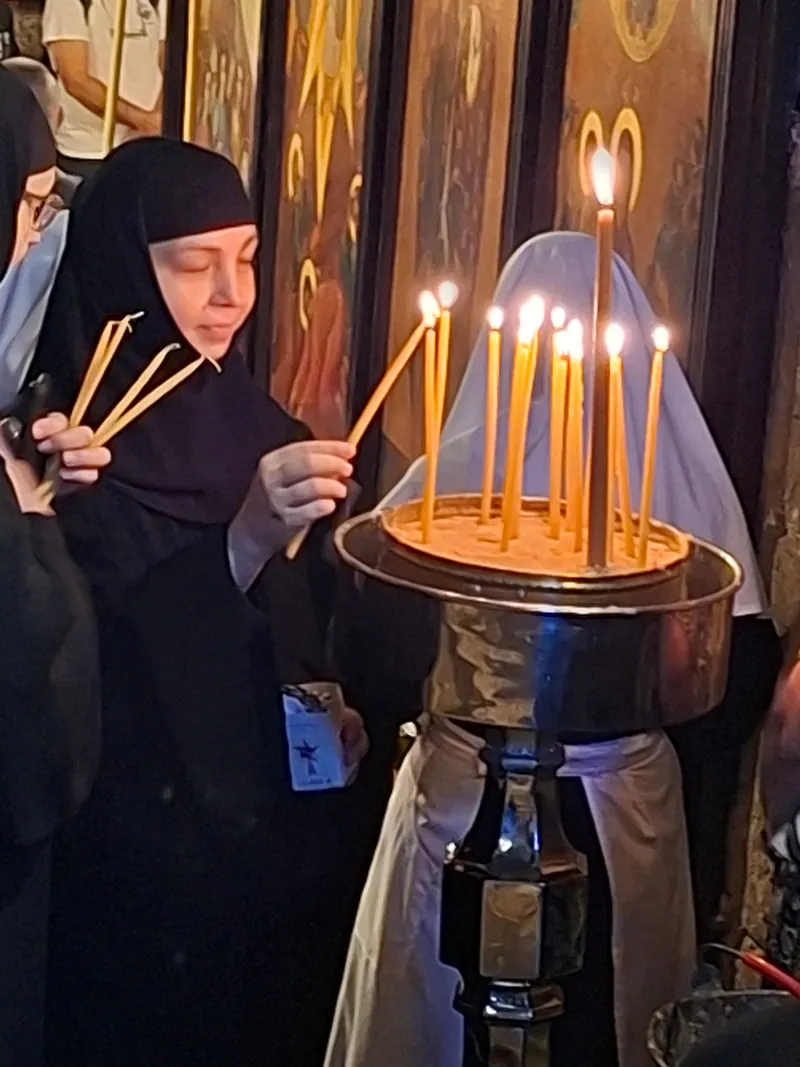 נזירה בלבוש שחור מדליקה נרות שעווה דקים במעמד נרות, לפני איקונות מוזהבות.