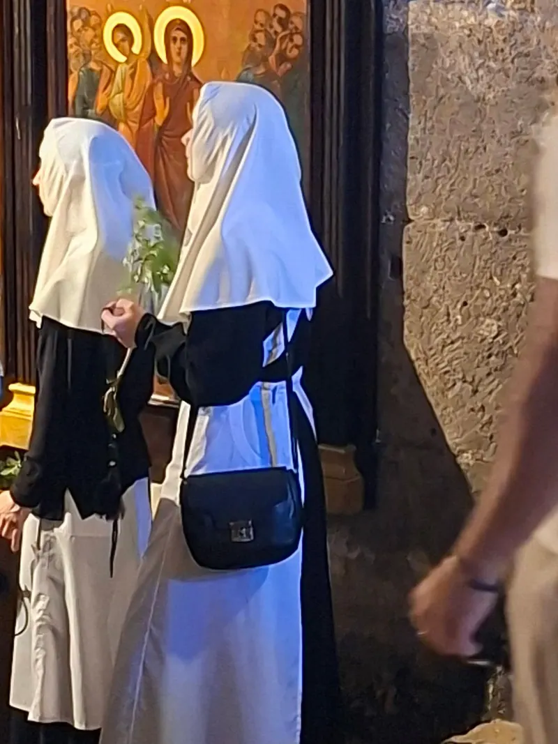 שתי נזירות בכיסויי ראש לבנים ובגלימות שחור־לבן אוחזות ענפים ירוקים לפני איקונה.