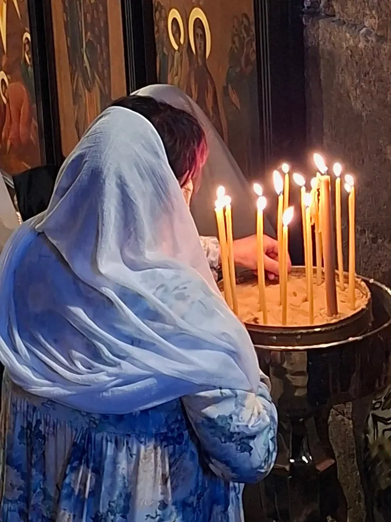 אישה בכיסוי ראש לבן־כחול מדליקה נרות שעווה דקים במעמד מלא חול, לפני איקונות.
