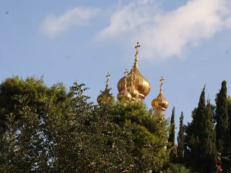 כיפות הבצל המוזהבות של כנסיית מריה מגדלנה מתנשאות מעל צמרות עצים וברושים, על רקע שמיים כחולים.