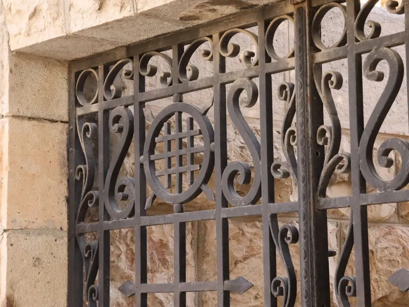 סורג ברזל מסולסל בחלון אבן, ובמרכזו צלב ירושלים בתוך עיגול.