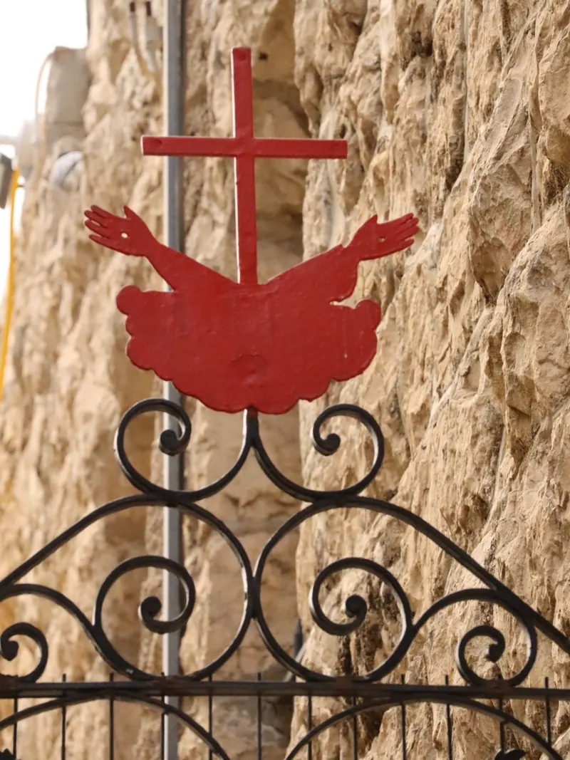 צלב מתכת אדום העולה מתוך ענן עם שתי זרועות פרושות, מעל שער ברזל מסולסל, על רקע קיר אבן מחוספס.