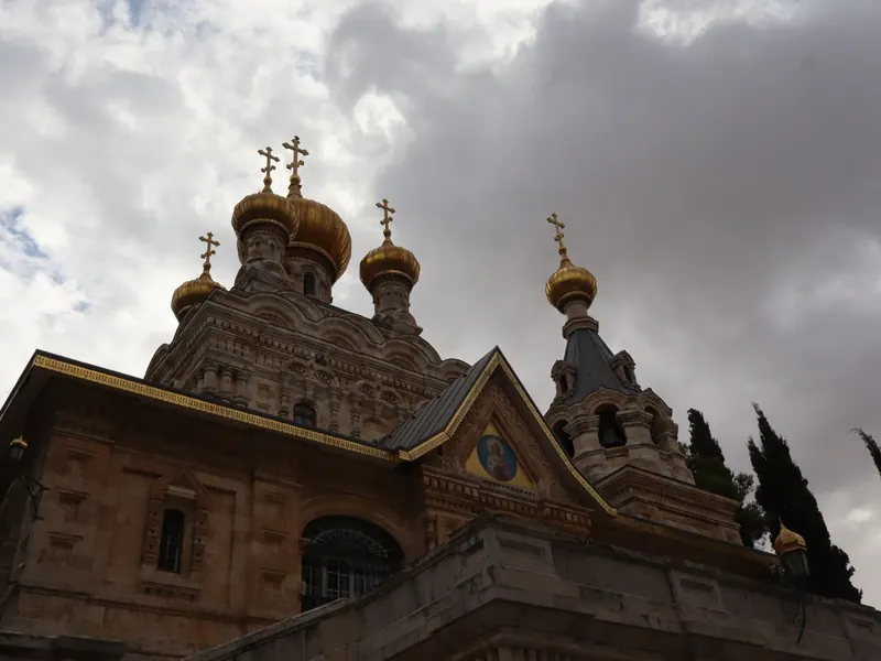 כנסיית מריה מגדלנה במבט מלמטה: חזית אבן, איקונין בגמלון וכיפות בצל מוזהבות תחת שמיים מעוננים.