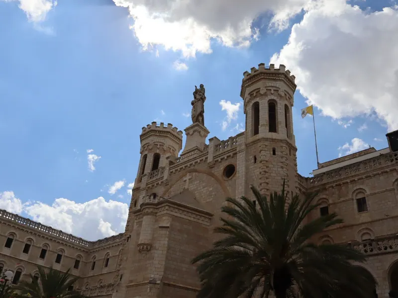 מבנה נוטרדאם של ירושלים: מגדלים משוננים ובראשם פסל מריה והתינוק, על רקע שמיים כחולים ועננים, ועצי דקל בחזית.