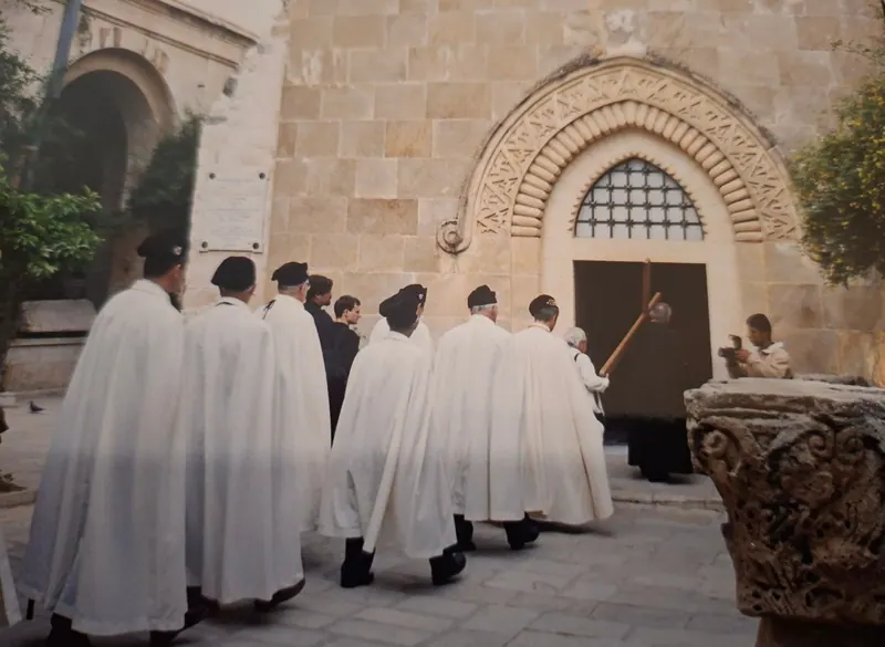 תהלוכת יום שישי בדרך הייסורים: משתתפים בגלימות לבנות ארוכות ובכומתות שחורות חוצים חצר מרוצפת אל פתח כנסייה תחת קשת אבן מגולפת; בראשם נושאים צלב עץ גדול.